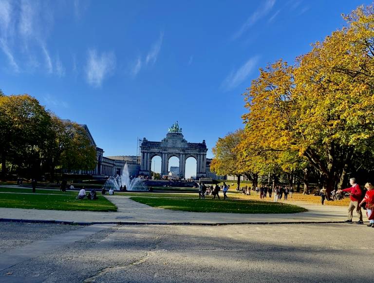 Park scene with a prominent archway in the background. Trees with autumn foliage line a wide path, and people stroll around a fountain under a clear blue sky.