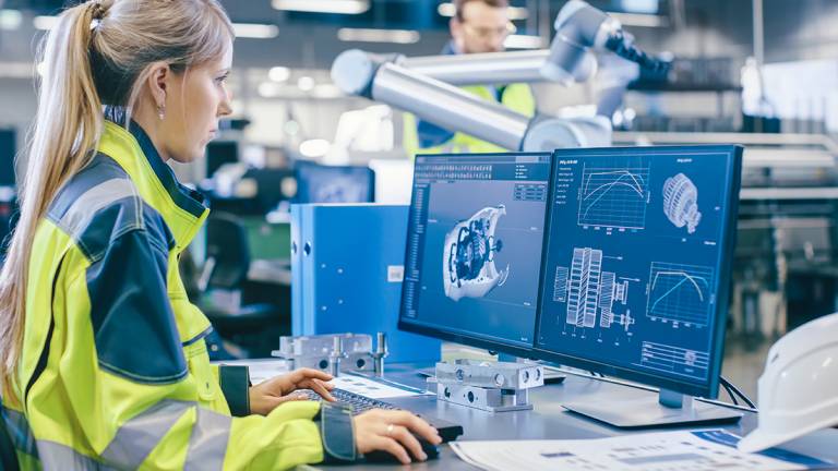 Tehtaalla: Naispuolinen koneinsinööri suunnittelee 3D-moottoria tietokoneellaan, miespuolinen automaatioinsinööri käyttää kannettavaa tietokonetta robottikäsivarren ohjelmointiin.