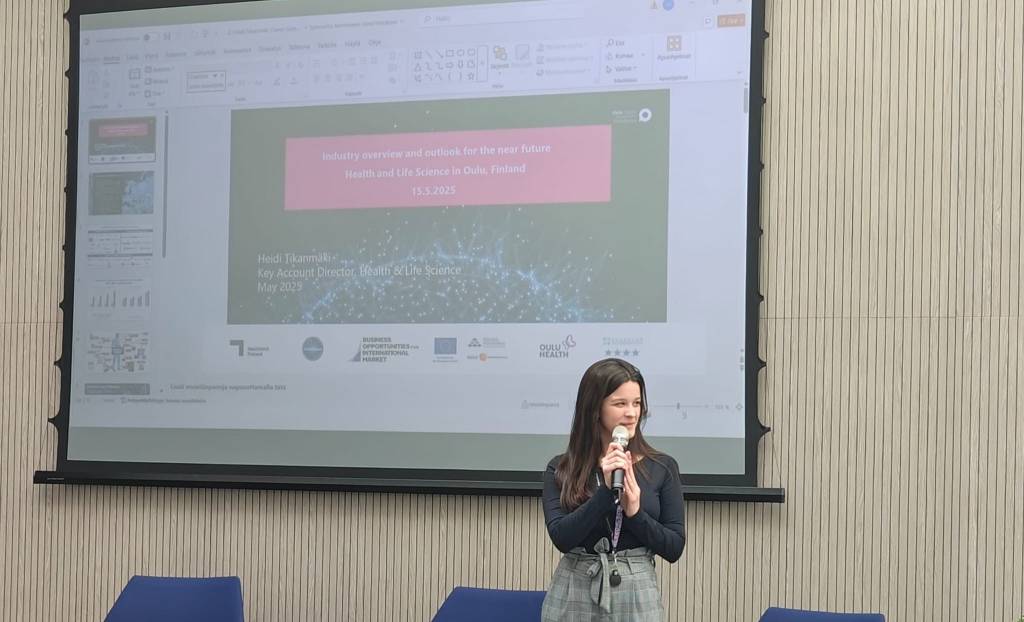 Beatriz with a microphone in her hand is standing in-front of a projector.