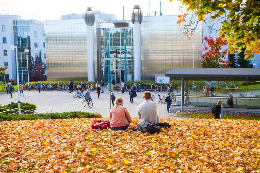Two students sitting on golden autumn leaves overlooking Tampere University city centre campus with modern glass buildings and bicycles.
