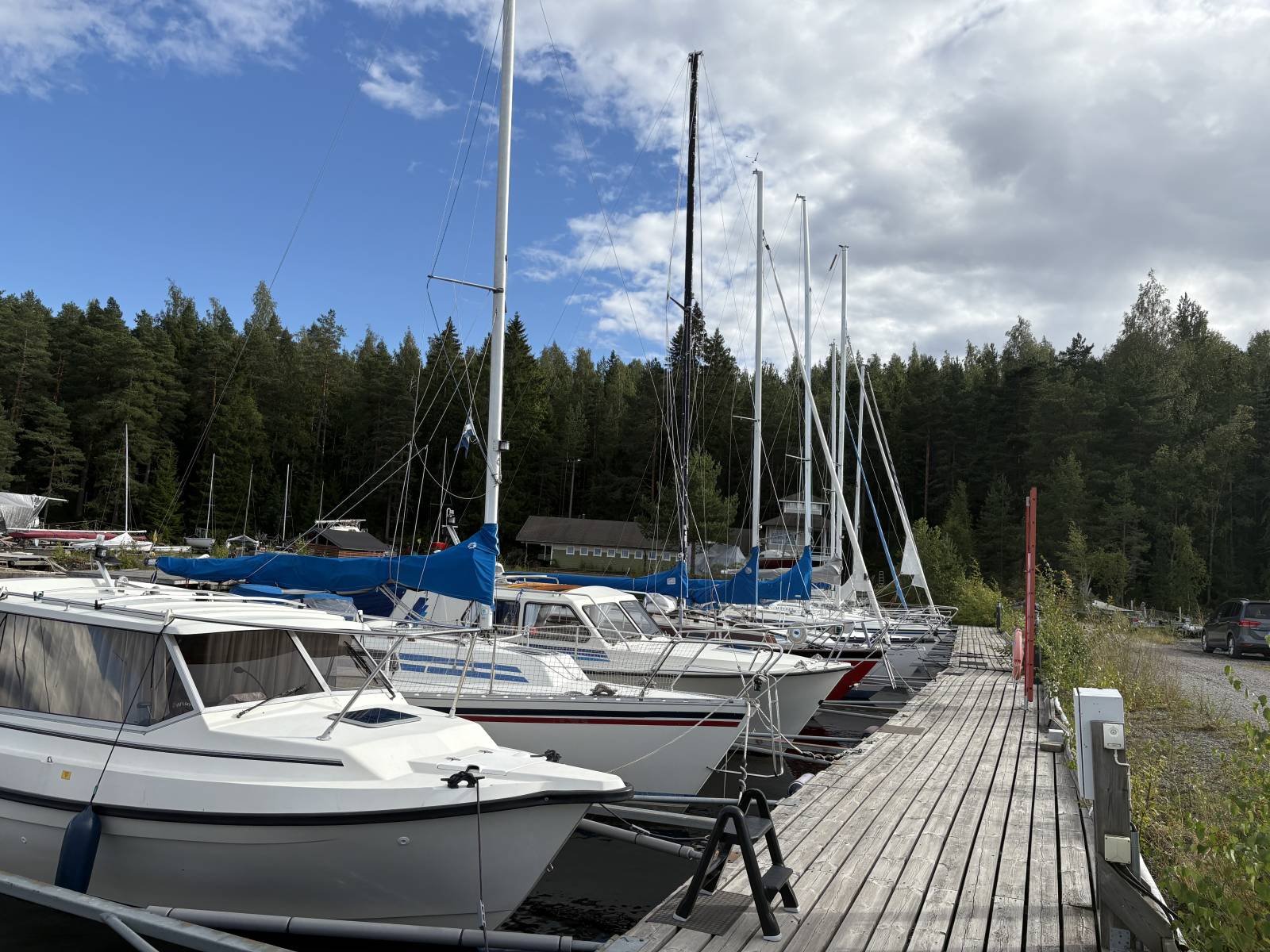 Marina dock with several sailboats moored along a wooden pier, backed by dense evergreen forest under a partly cloudy sky in Rauhaniemi, Tampere.