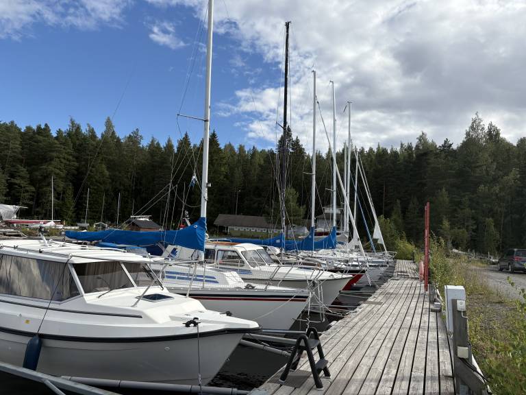 Marina dock with several sailboats moored along a wooden pier, backed by dense evergreen forest under a partly cloudy sky in Rauhaniemi, Tampere.