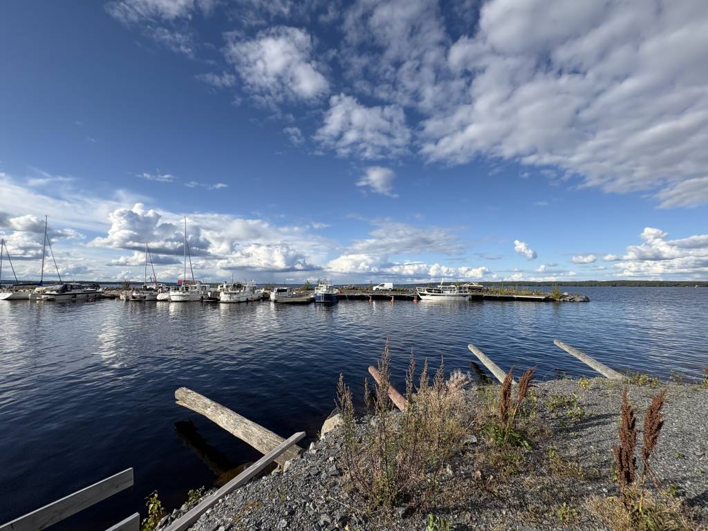 Scenic view of a marina in Rauhaniemi, Tampere, with boats moored at wooden docks on calm blue water, surrounded by rocky shoreline with sparse vegetation under a dramatic sky with white clouds.