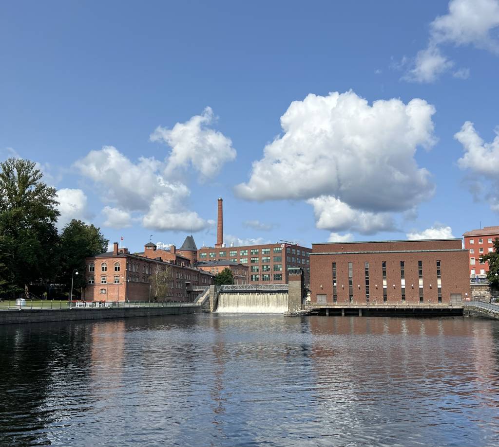 Historic red brick industrial buildings along the Tammerkoski rapids in Tampere, featuring a tall chimney, waterfall, and their reflections in the calm water under a blue sky with white clouds.
