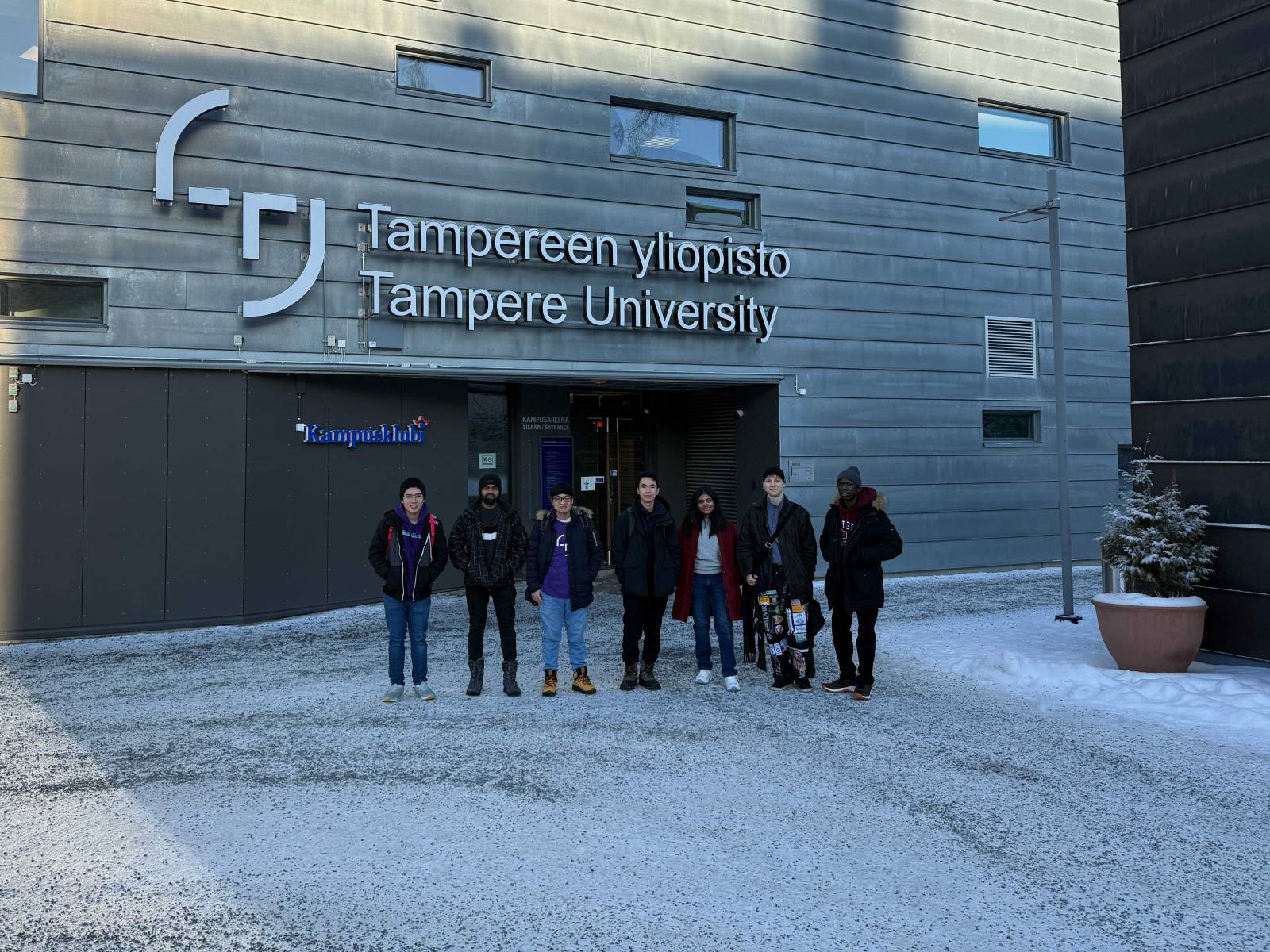 Seven students standing in front of Tampere University's main entrance building in winter with snow on the ground.