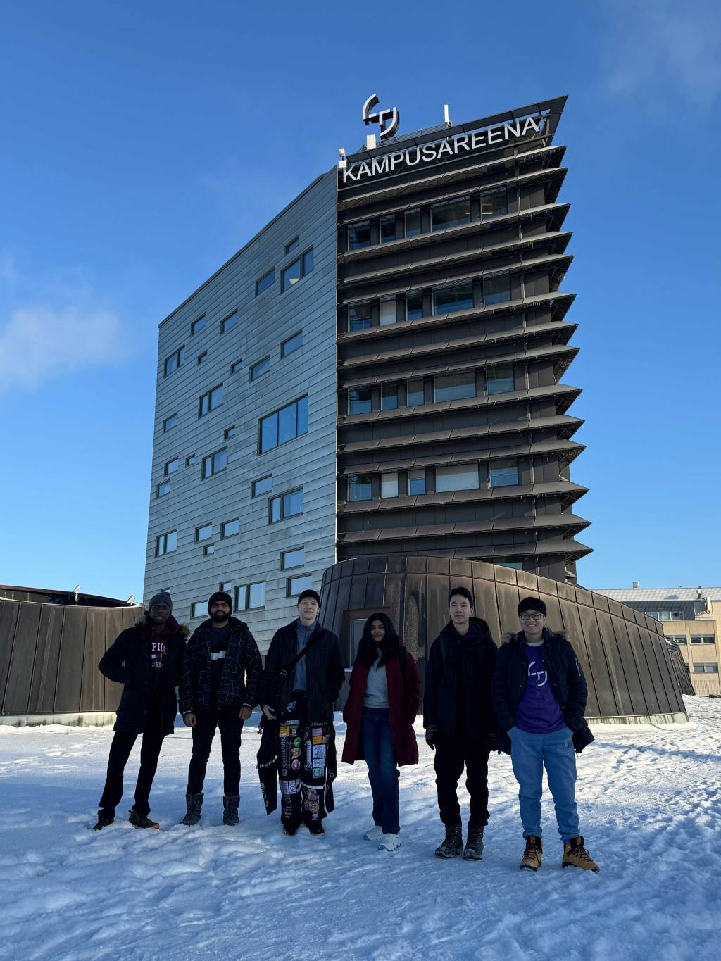 Six international students standing in the snow in front of the Kampusareena tower building with clear blue sky.