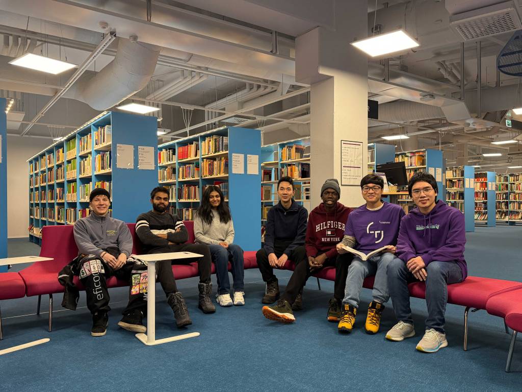 Group of seven international students sitting together in a modern library with blue bookshelves at Tampere University.
