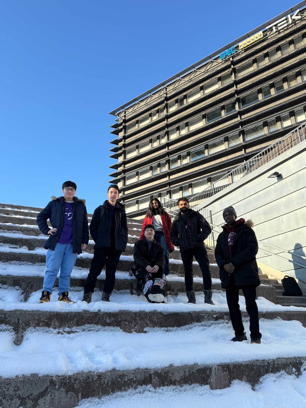 Six international students standing on snowy steps in front of the Kampusareena building at Tampere University.