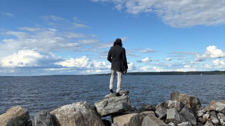Person standing on rocky shoreline overlooking Lake Näsijärvi in Rauhaniemi, Tampere, with dramatic clouds and forested coastline in the background.