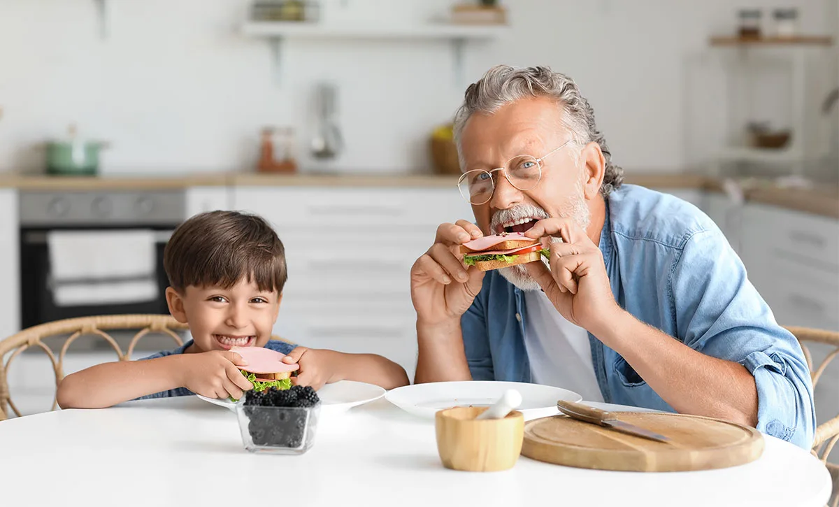Little boy with his grandfather eating tasty sandwiches at table in kitchen