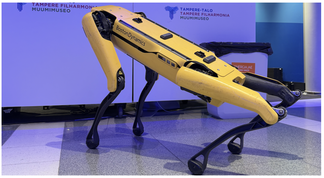 A close-up side view of the Boston Dynamics Spot robot standing in a lit event space, showing its yellow body and legs