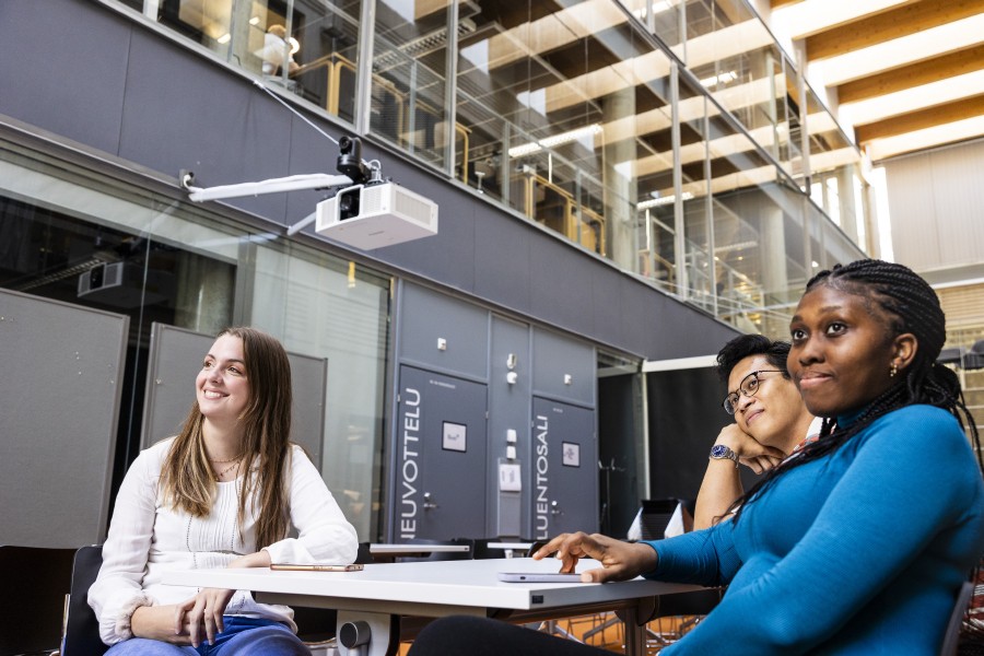 Kolme opiskelijaa istuu pöydän ääressä, katsovat ohi kamerasta.