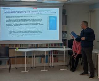 Image shows Dr Kaunisto standing near the white screen and holding a book