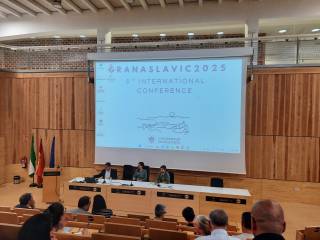 The image shows a conference auditorium with three speakers at a panel table, a slide with the conference logo behind them