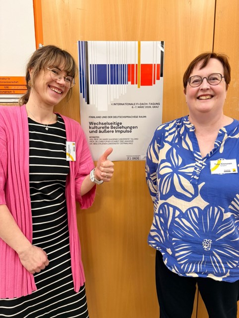 Maija Hirvonen and Tuija Kinnunen smile at the camera in front of the conference poster. Maija Hirvonen shows thumb up.
