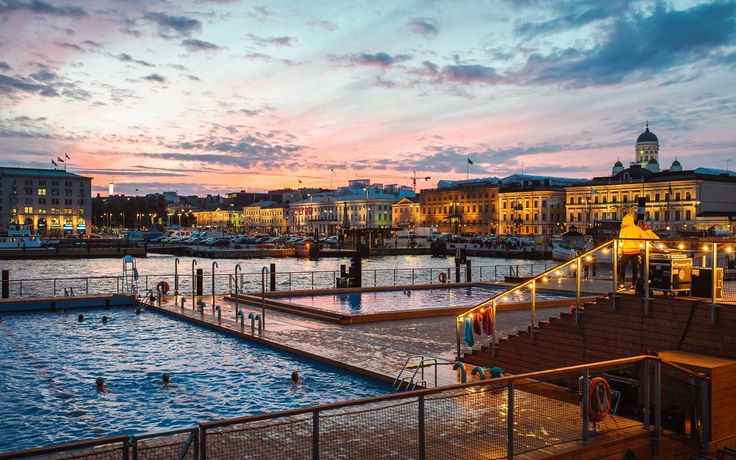 An outdoor public pool and sauna complex on the Helsinki waterfront at dusk, with the illuminated city skyline and Helsinki Cathedral in the background.