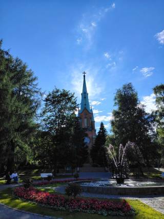 A church tower with a teal spire rises above green trees and a flowering park with a fountain on a sunny summer day in Tampere, Finland.