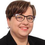 Hymyilevä tummahiuksinen, silmälasipäinen nainen katsoo kameraan.