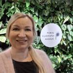 Hymyilevä vaaleahiuksinen nainen seisoo viherseinän edessä. Seinässä on kyltti, jossa lukee porin yliopistokeskus.