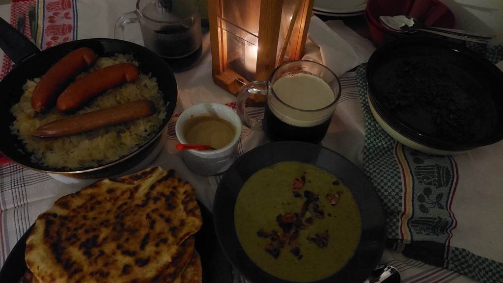 Foods on a table, over cloths. Foods include sausages, greenish soup, flatbreads, chocolate mousse, mustard and beer. In the middle of the table is a lantern, with candle burning inside.