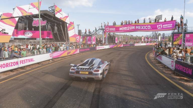 A silver-gray car driving on the road in a middle of a festival with a stage in a background