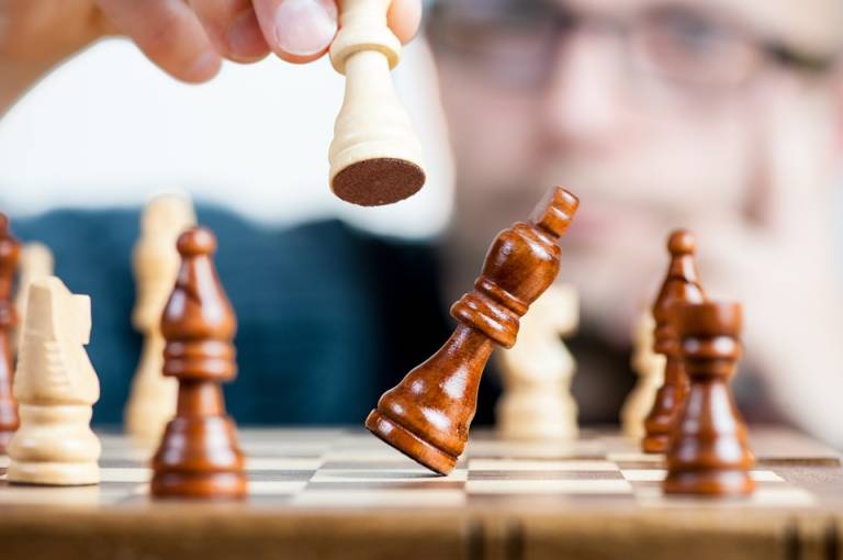 Image is focused on a chessboard with white and brown pieces and in the middle of it is one brown piece falling over after a person knocked it over with a white piece.