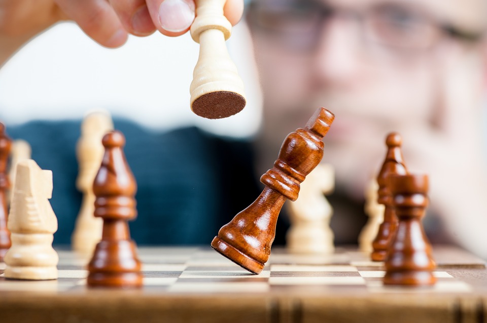 Image is focused on a chessboard with white and brown pieces and in the middle of it is one brown piece falling over after a person knocked it over with a white piece.