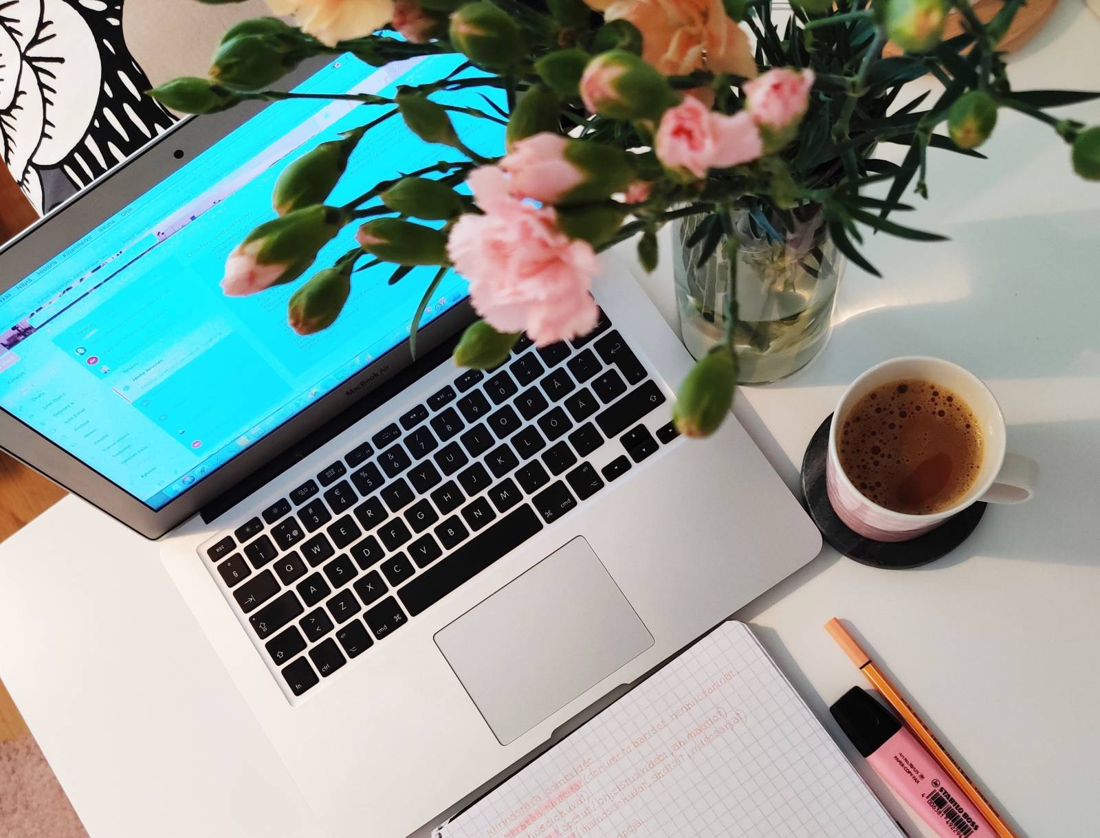 Kirjoituspöydällä värikäs kukkakimppu, kuppi kahvia, muistiinpanovihko, kyniä sekä kannettava tietokone.