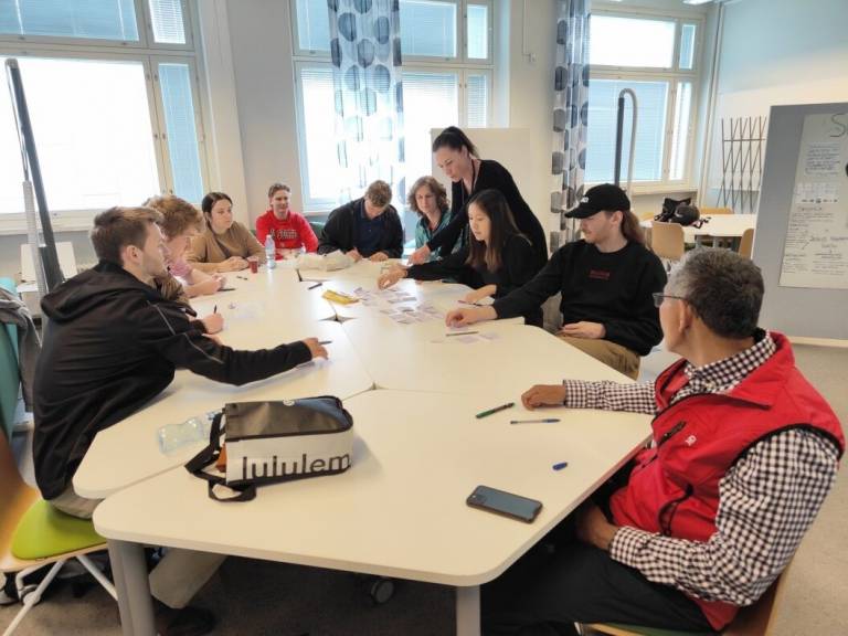 Opiskelijaryhmä ja ohjaaja istuvat työpajassa pöydän ääressä.