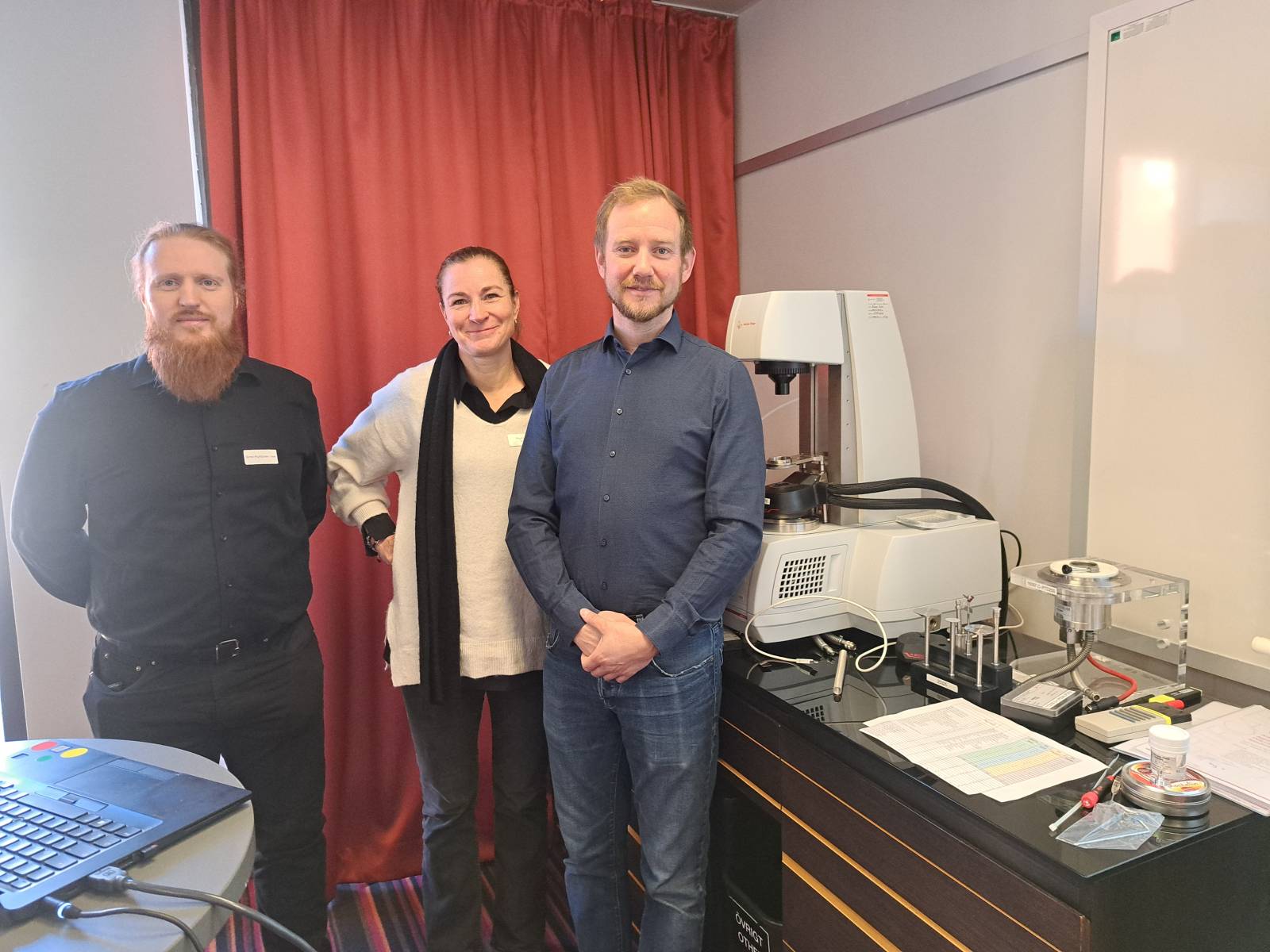 Three people standing, table with machines on it in the background.